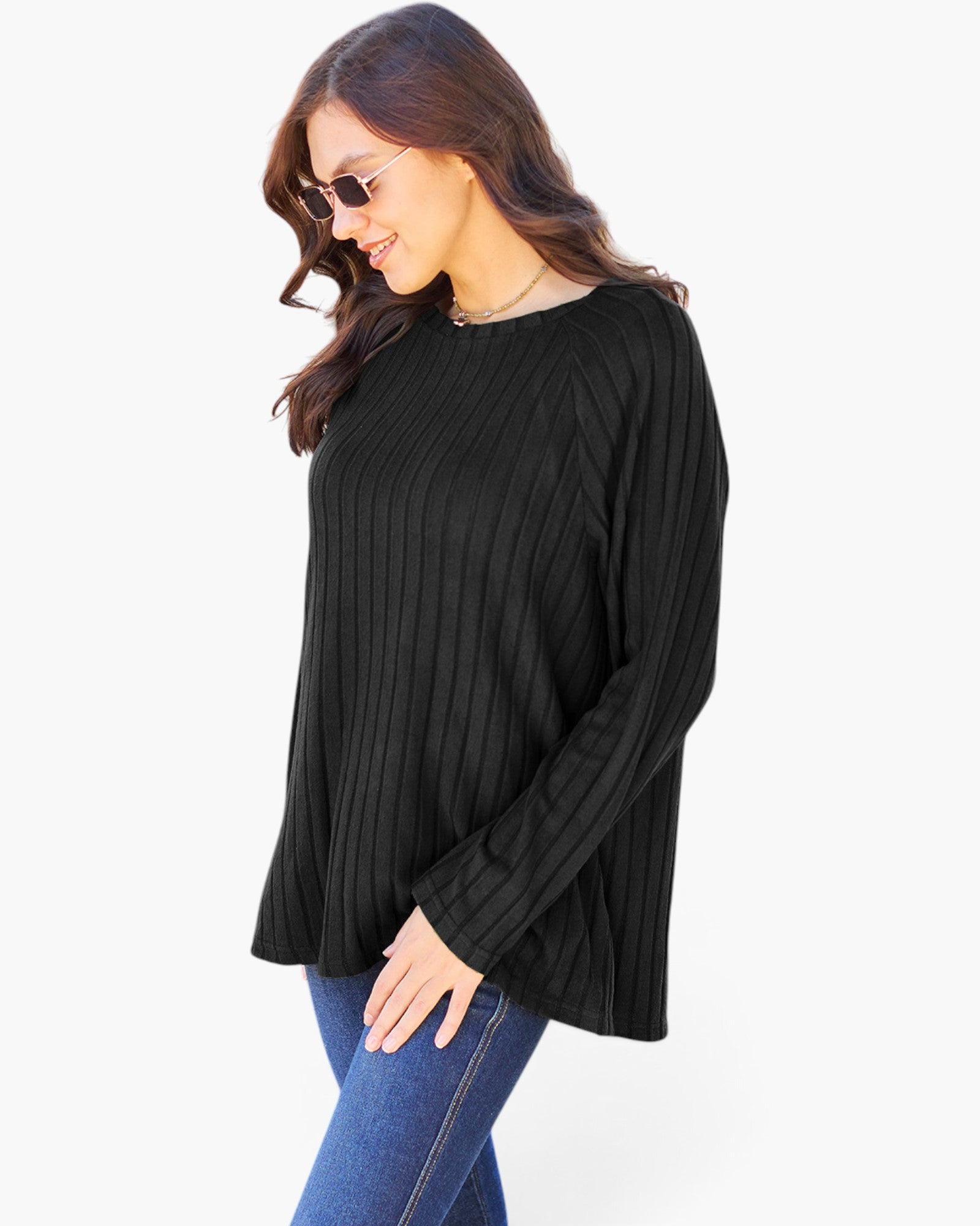 A woman wearing a black ribbed long-sleeve top and blue jeans, photographed against a plain light gray studio background.