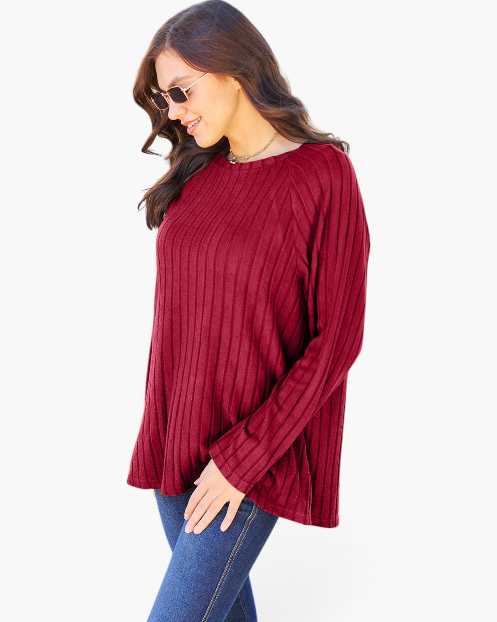 A woman wearing a ribbed long-sleeve knit top in deep burgundy red, paired with blue jeans and sunglasses, shown against a plain white background.