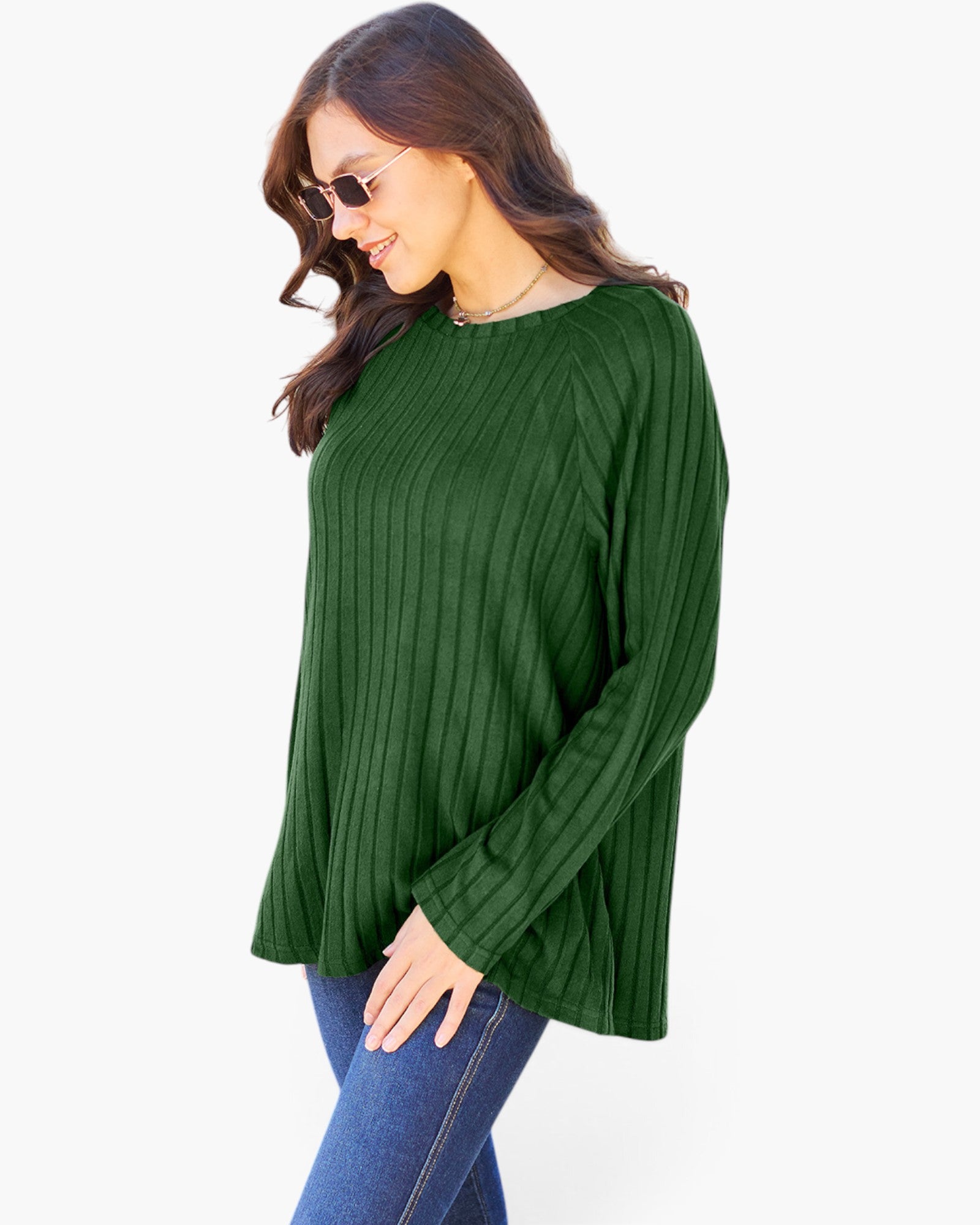 A woman wearing a dark green ribbed knit long-sleeve top and blue jeans, photographed against a plain light gray studio background.