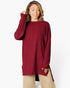 A woman wearing a ribbed knit long-sleeve tunic sweater in deep burgundy, paired with beige pants, against a plain white studio background.