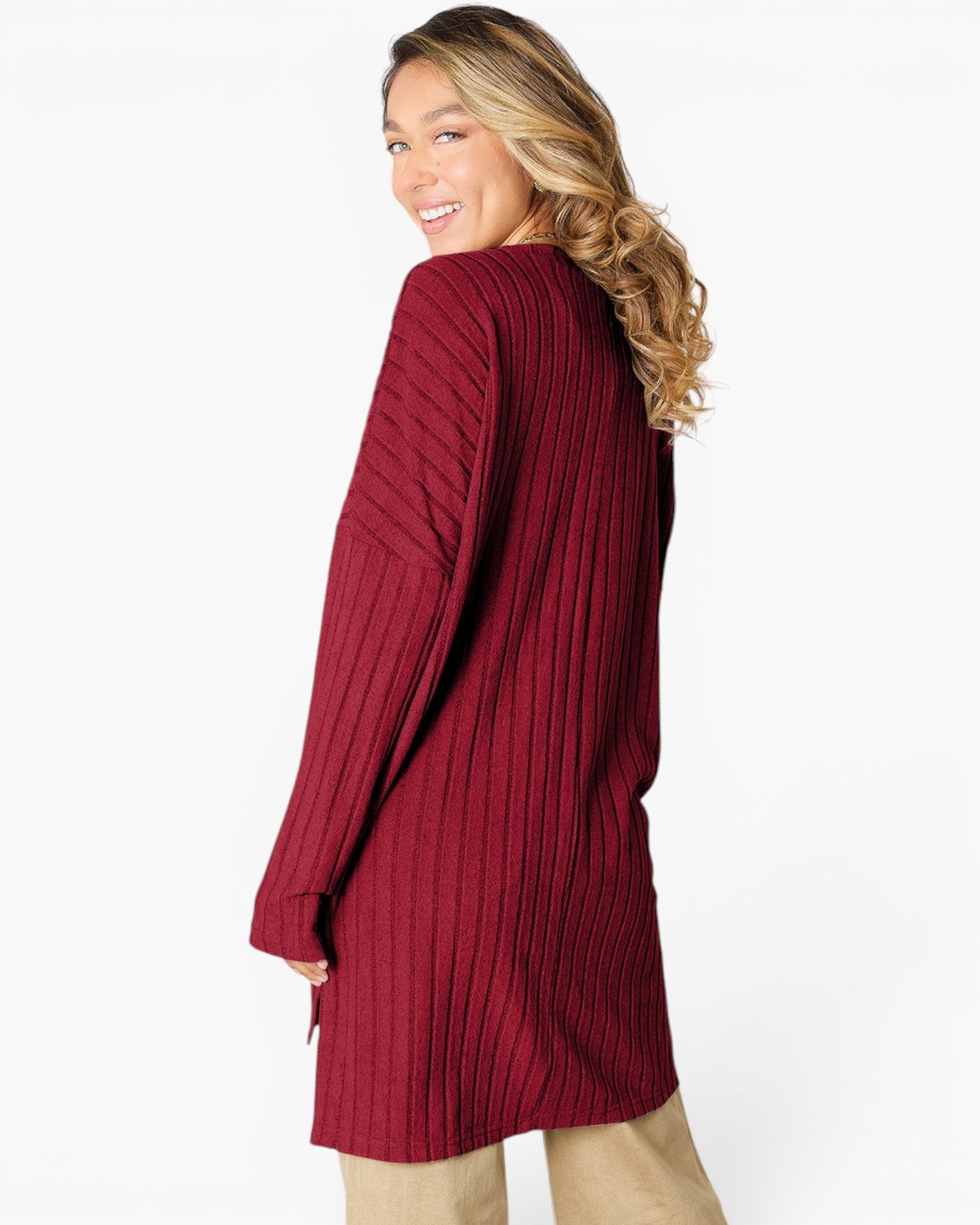 A woman wearing a ribbed knit long-sleeve tunic sweater in burgundy, shown from a side-back angle against a plain white studio background.