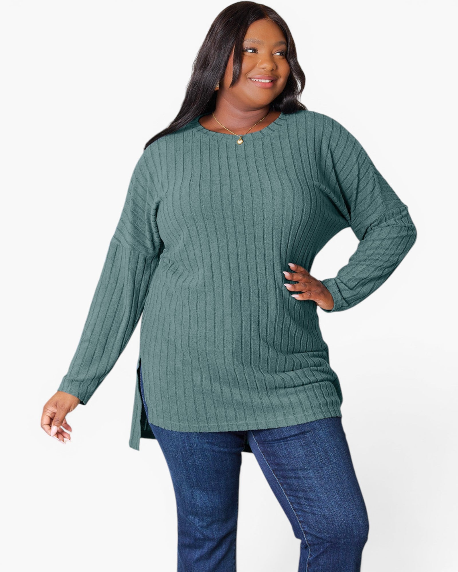 A woman modeling a ribbed knit long-sleeve tunic top in sage green, paired with blue jeans, against a plain white studio background.