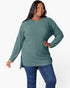 A woman modeling a ribbed knit long-sleeve tunic top in sage green, paired with blue jeans, against a plain white studio background.