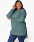 A woman modeling a ribbed knit long-sleeve tunic top in sage green, paired with blue jeans, against a plain white studio background.
