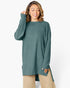 A woman wearing a ribbed knit long-sleeve tunic sweater in a muted teal green, paired with beige pants, against a plain white studio background.