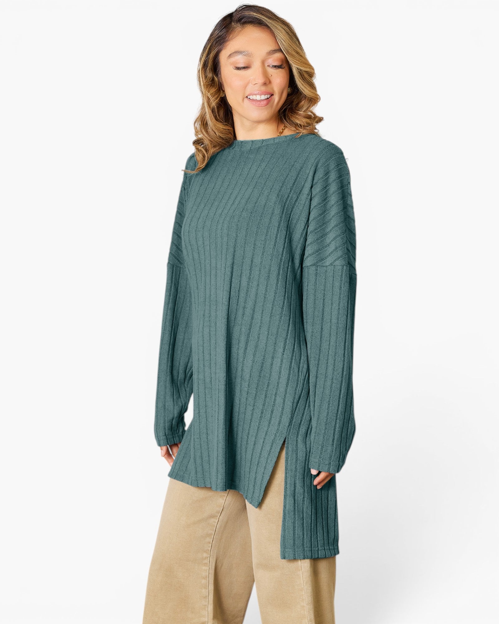 A woman wearing a ribbed knit long-sleeve tunic top in a muted teal green color, paired with beige pants, against a plain white studio background.