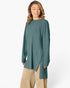 A woman wearing a ribbed knit long-sleeve tunic top in a muted teal green color, paired with beige pants, against a plain white studio background.