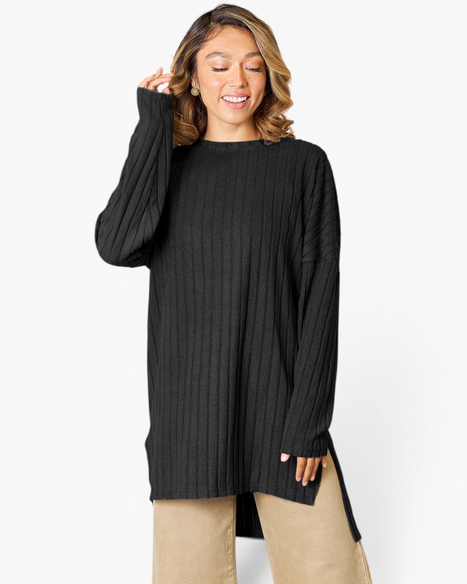 A woman wearing a black ribbed long-sleeve tunic sweater with a high-low hem and side slits, paired with beige pants, against a plain white studio background.