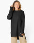 A woman wearing a black ribbed long-sleeve tunic sweater with a high-low hem and side slits, paired with beige pants, against a plain white studio background.