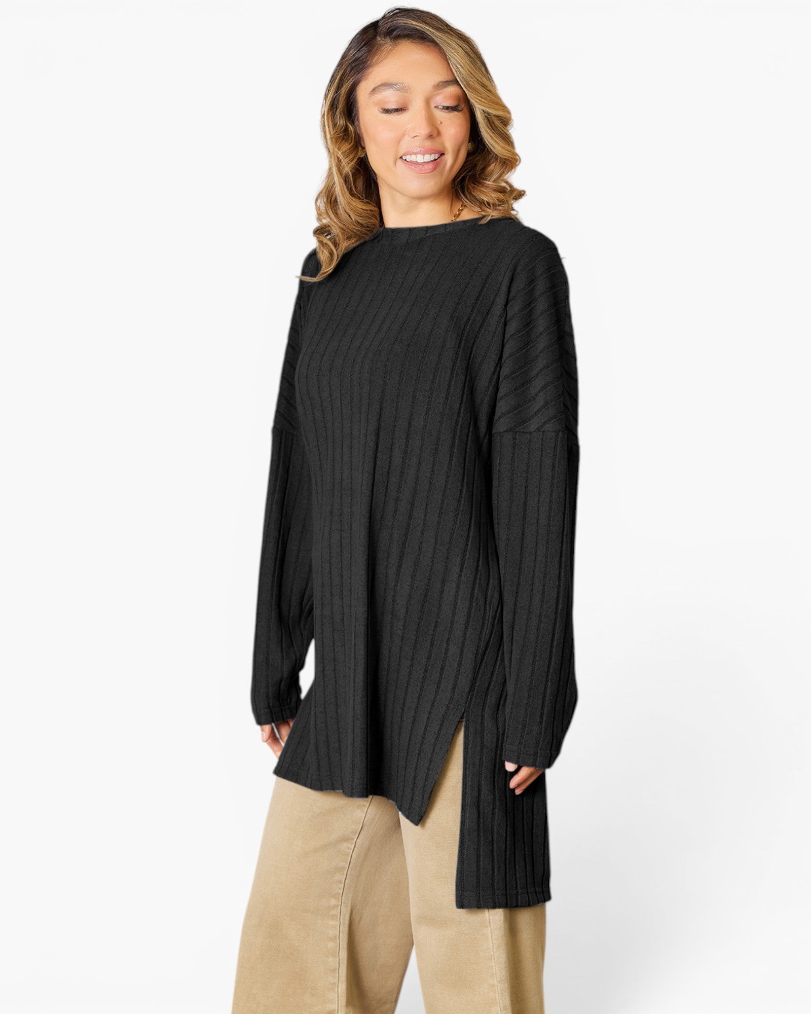 A woman wearing a black ribbed long-sleeve tunic top with a high-low hem and side slits, paired with beige pants, against a plain white studio background.
