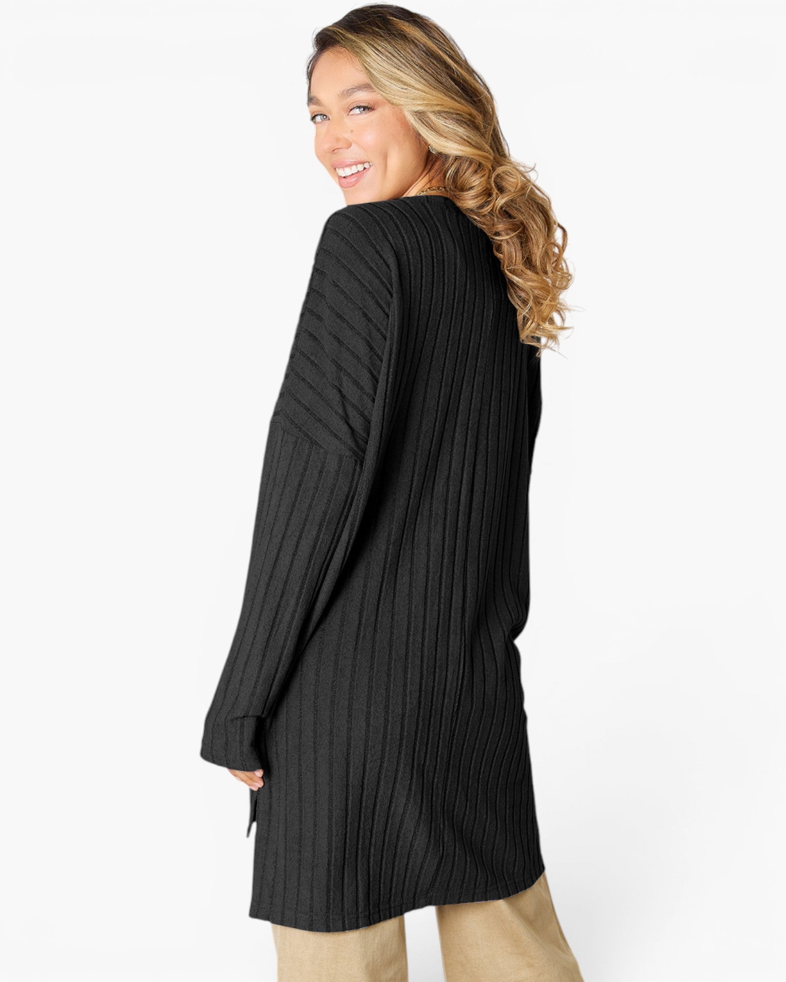 A woman wearing a black ribbed knit long-sleeve tunic sweater, shown from a side-back angle against a plain white studio background.