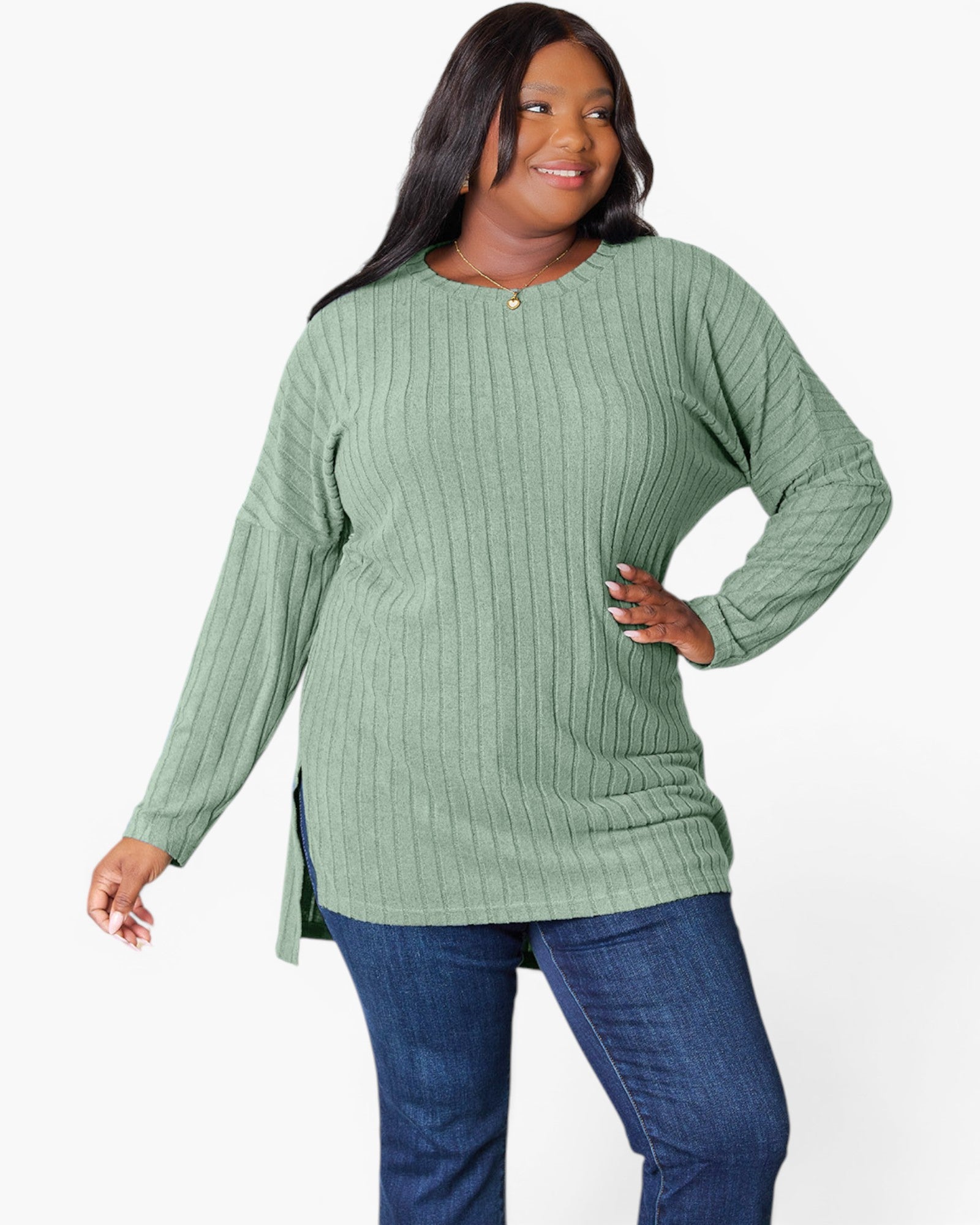 A plus-size woman wearing a sage green ribbed knit long-sleeve tunic top with blue jeans, posing against a solid white studio background.