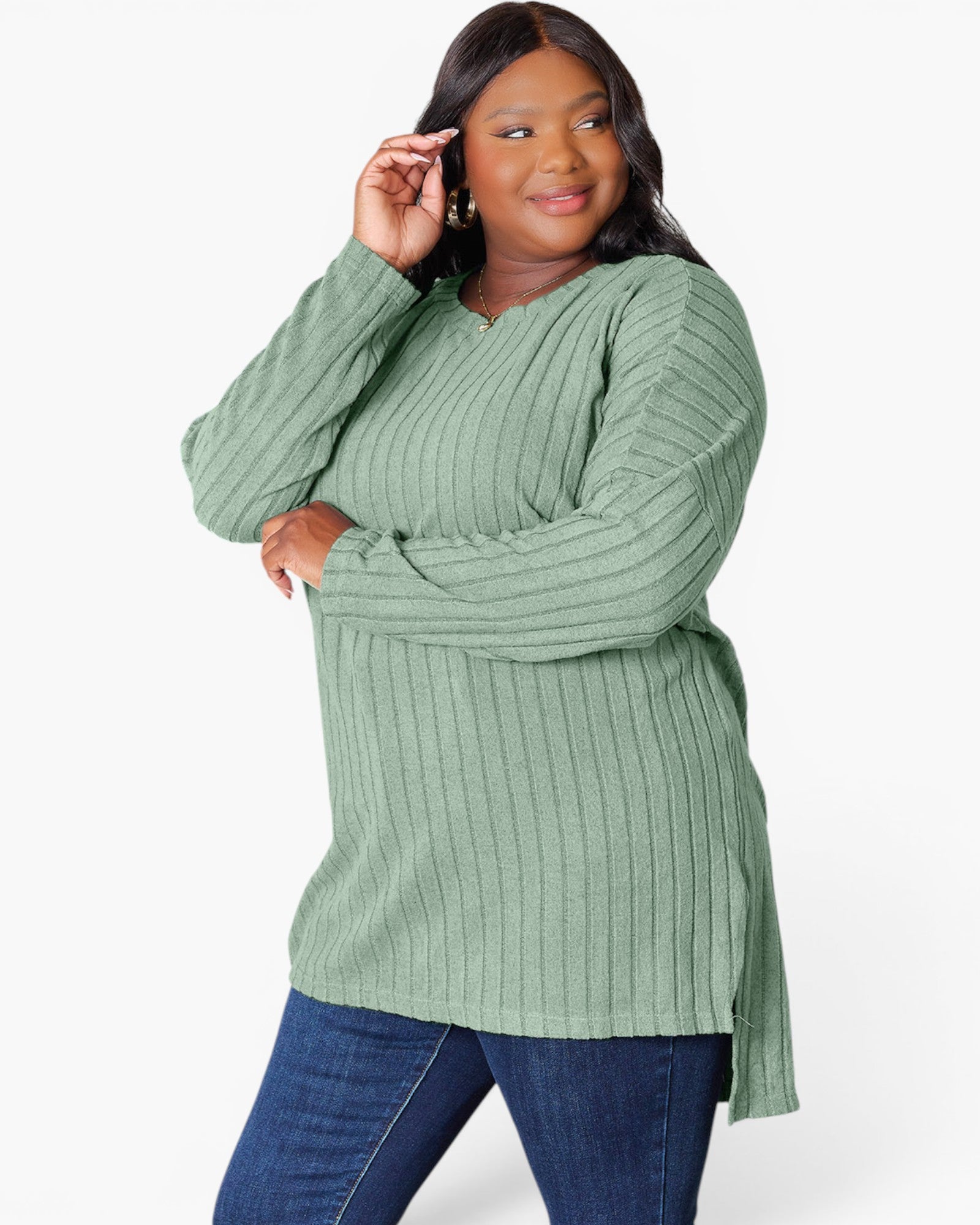 A woman modeling a sage green ribbed knit long-sleeve tunic top with blue jeans, photographed against a clean white studio background.
