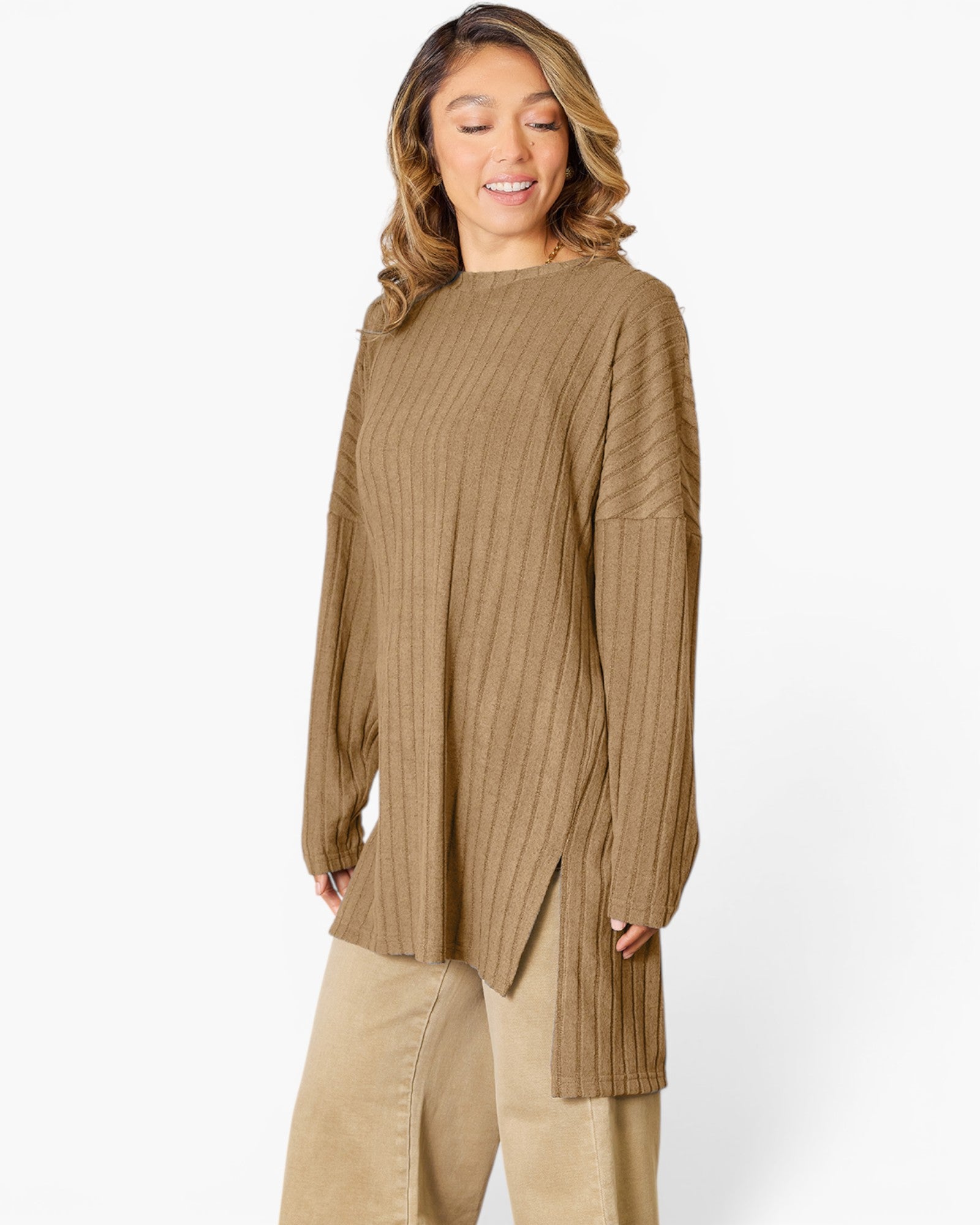 A woman wearing a brown ribbed knit long-sleeve tunic top with a high neckline and side slit, paired with beige pants, against a light gray studio background.
