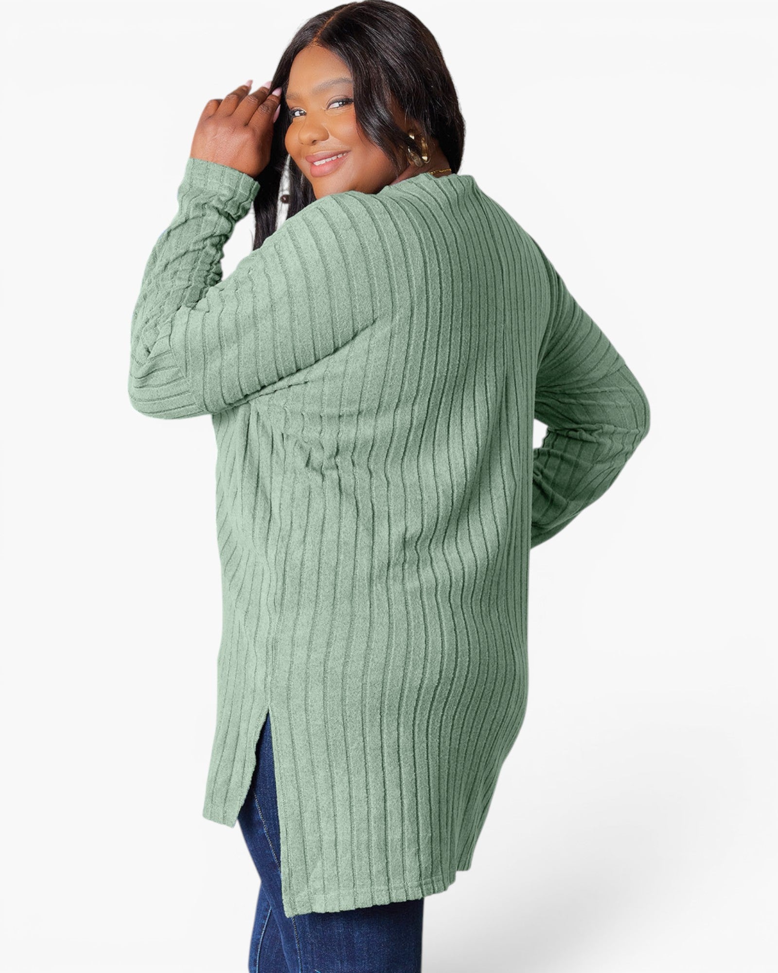 A woman wearing a sage green ribbed knit long-sleeve tunic sweater, shown from a three-quarters back view against a light gray studio background.
