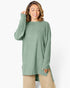 A woman wearing a sage green ribbed knit long-sleeve tunic top with beige pants, posing against a light gray studio background.