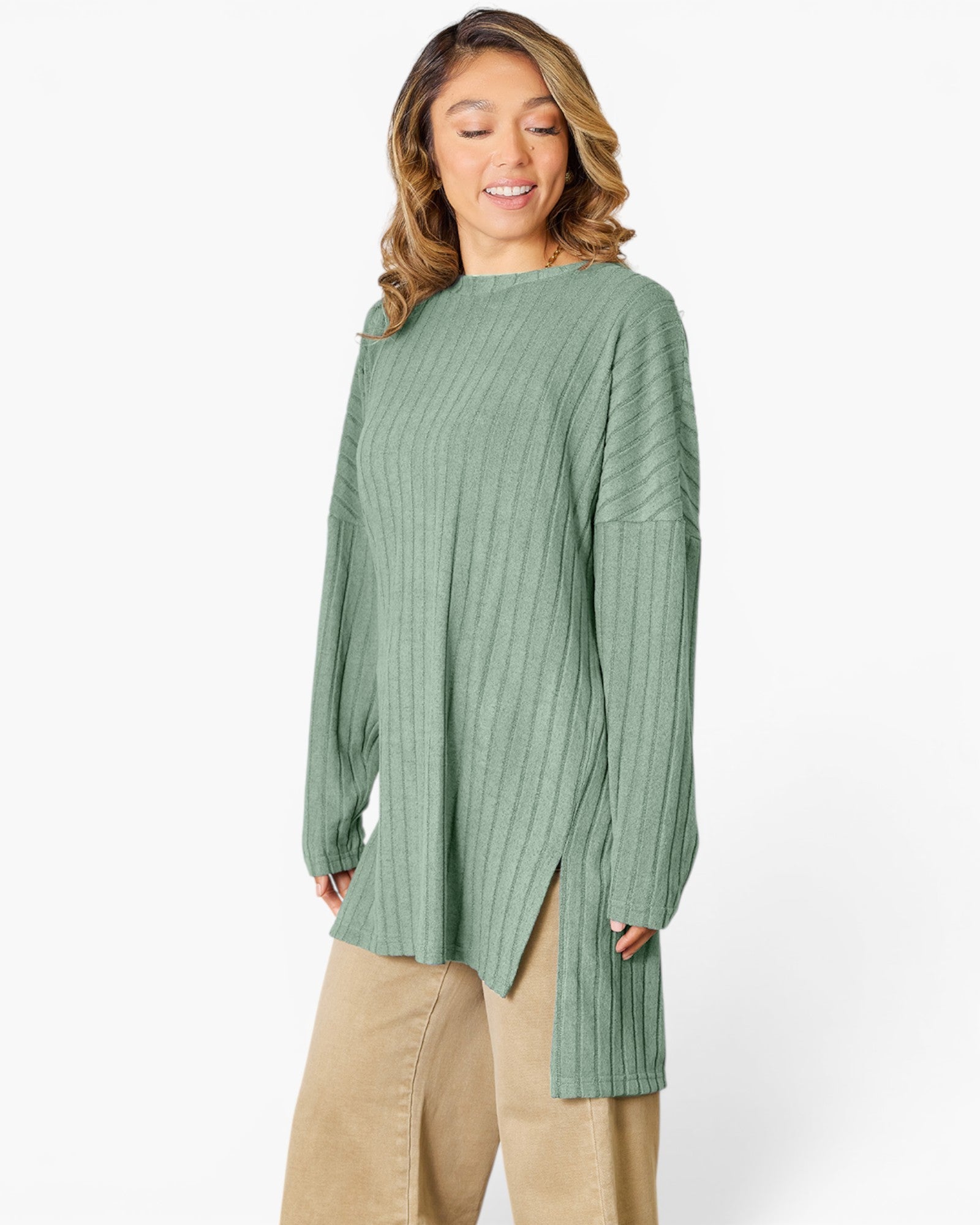 A woman wearing a sage green ribbed knit tunic sweater with a high neckline and side slits, paired with beige pants, against a plain white studio background.