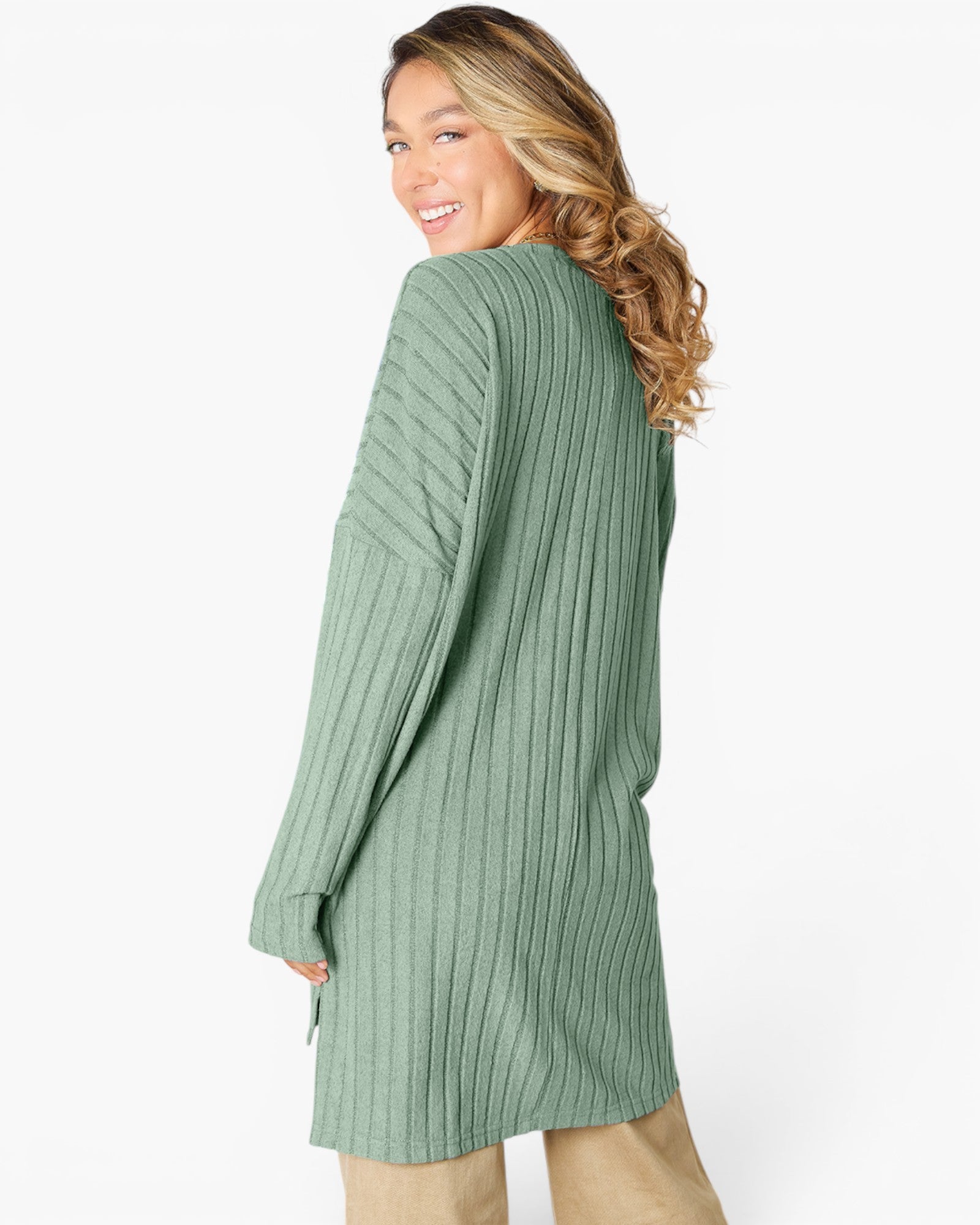 A woman wearing a sage green ribbed knit long-sleeve tunic sweater, shown from a side-back angle against a plain white studio background.