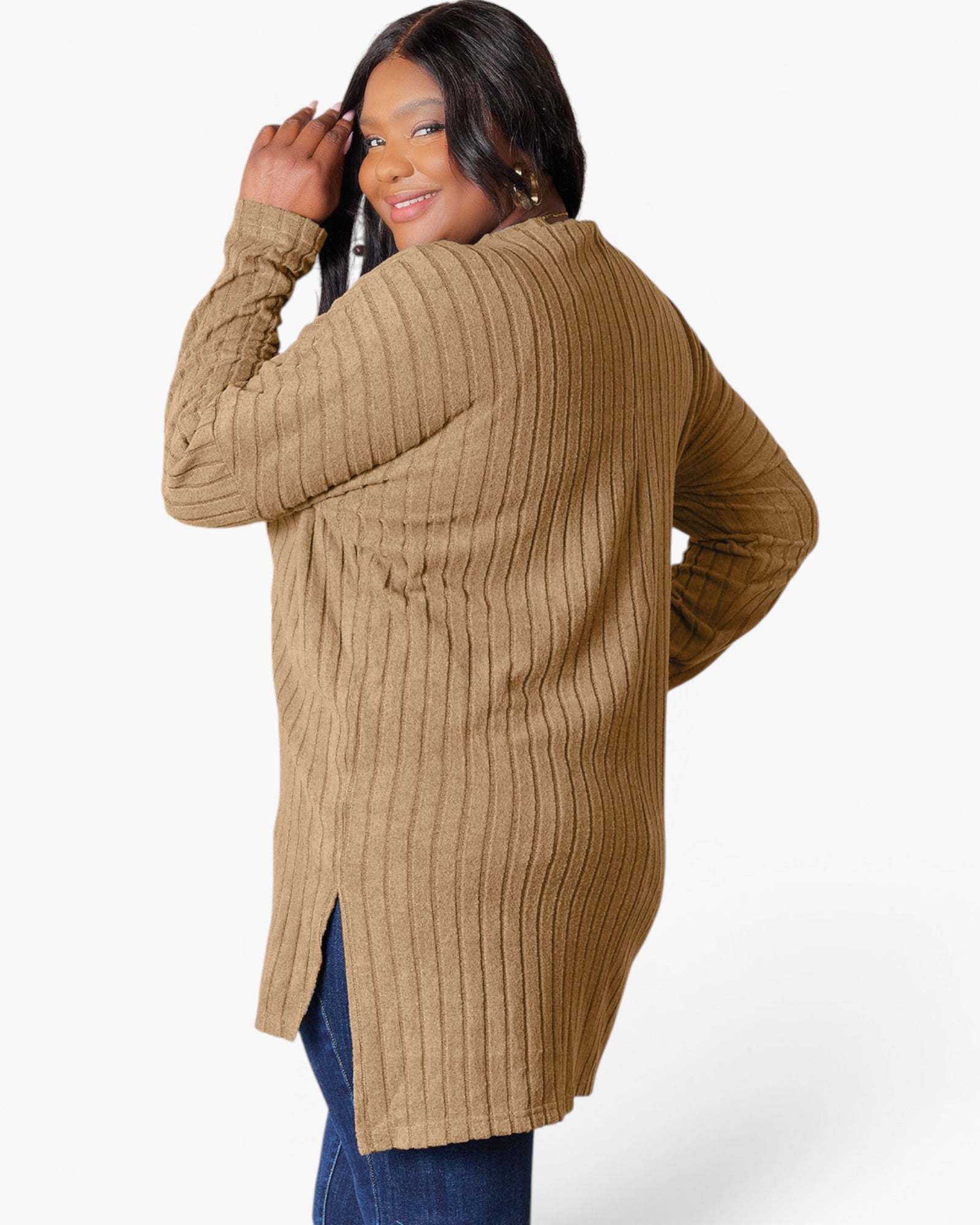 A woman wearing a camel-colored ribbed knit tunic sweater with blue jeans, photographed from the back against a clean white studio background.