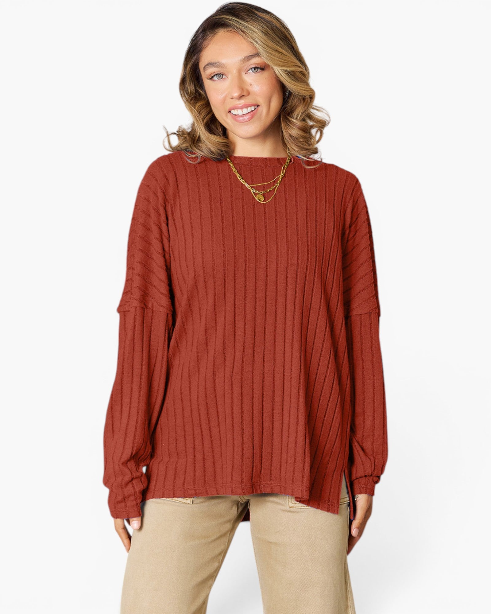 A woman wearing a rust-colored ribbed knit long-sleeve top and beige pants, standing against a plain white studio background.