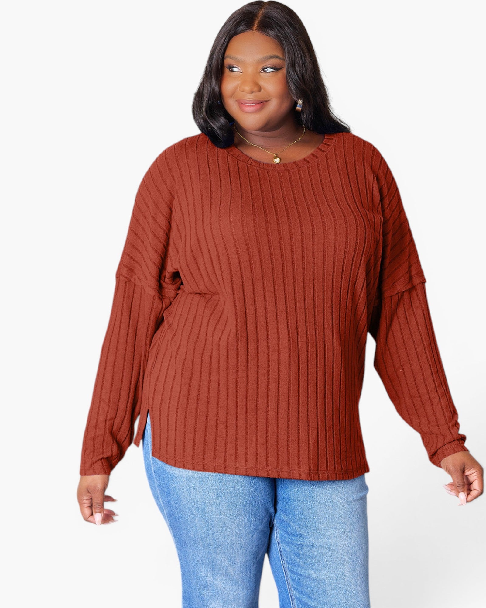 A woman wearing a rust-colored ribbed knit long-sleeve top and blue jeans, standing against a plain white background.