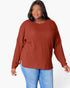 A woman wearing a rust-colored ribbed knit long-sleeve top and blue jeans, standing against a plain white background.