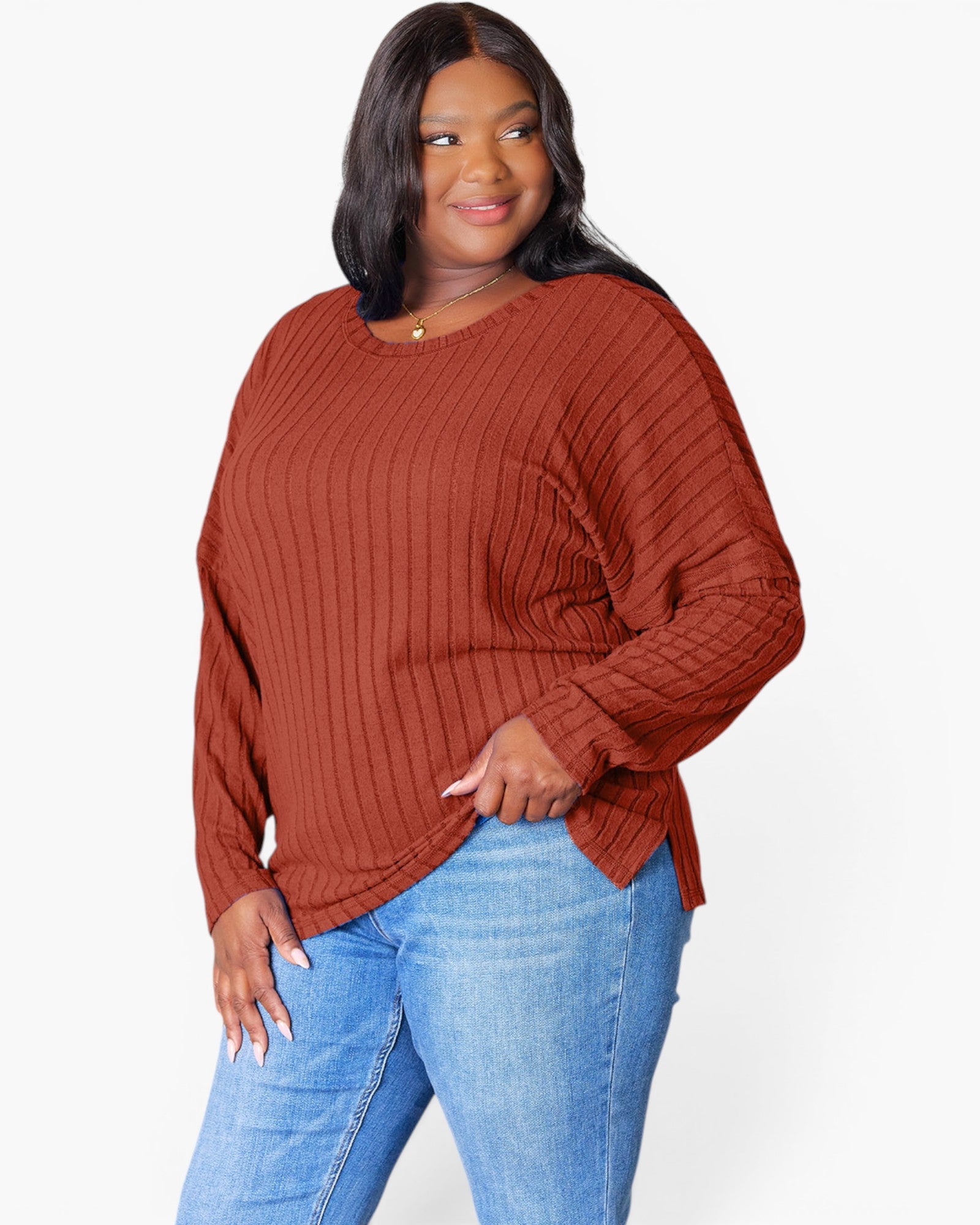 A woman modeling a rust-colored ribbed knit sweater with a boat neckline and long sleeves, paired with blue jeans, against a plain white background.
