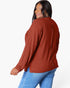 A woman wearing a rust-colored ribbed knit sweater and blue jeans, photographed from the back against a plain white studio background.