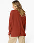 A woman shown from the back wearing a rust-colored ribbed knit sweater and beige pants against a plain white background.
