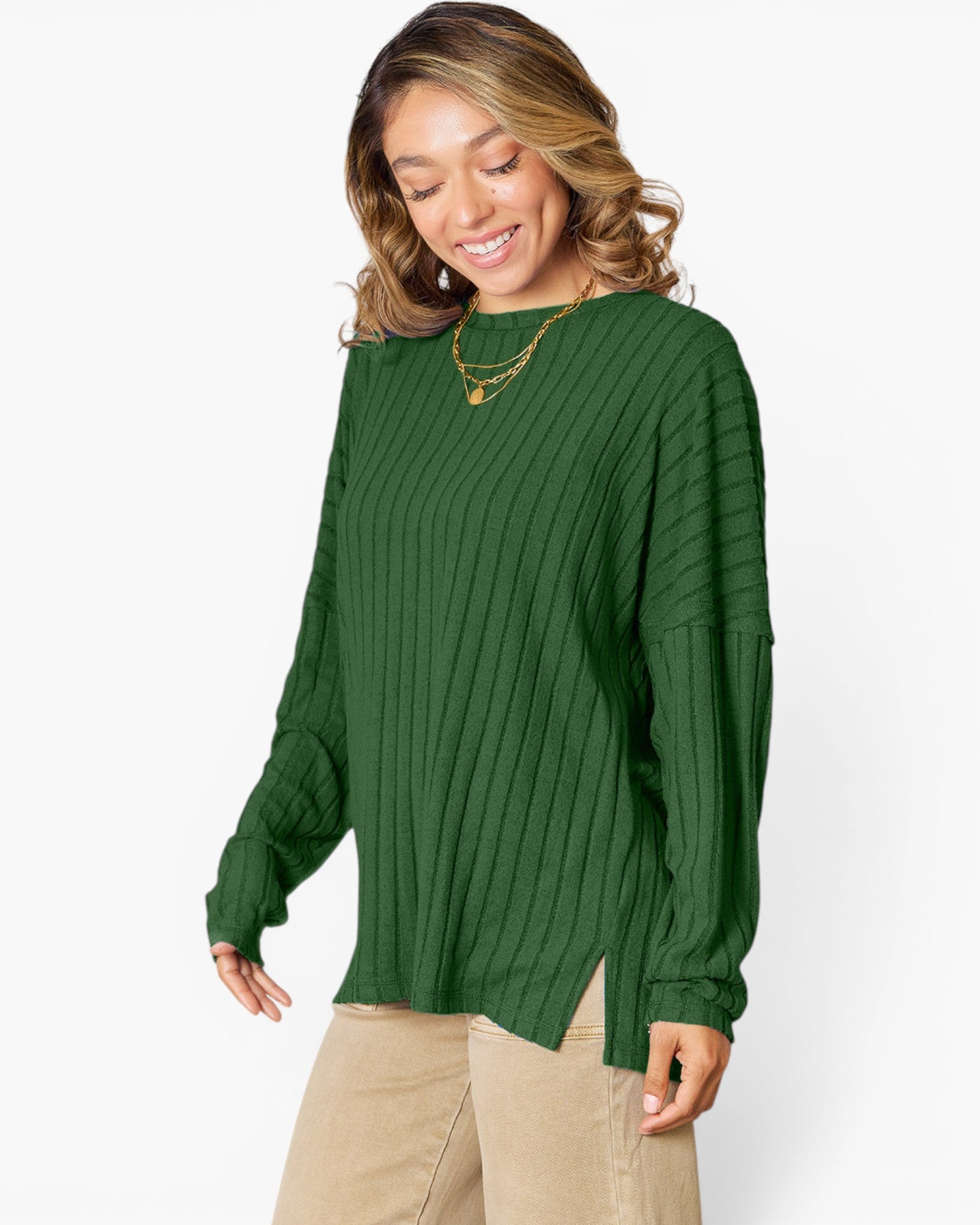 A woman wearing a dark green ribbed knit long-sleeve top with a crew neck and side slit hem, paired with beige pants, against a plain white background.