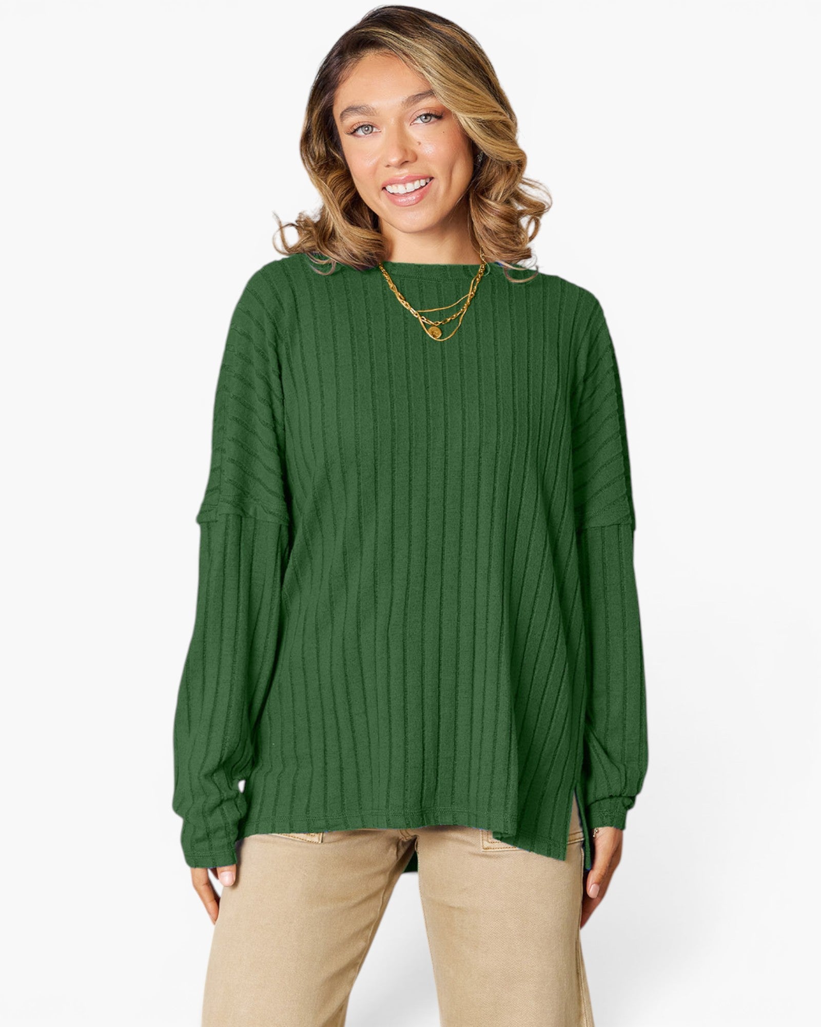 A woman wearing a forest green ribbed knit long-sleeve top with a boat neckline, paired with beige pants, against a plain white studio background.