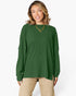 A woman wearing a forest green ribbed knit long-sleeve top with a boat neckline, paired with beige pants, against a plain white studio background.