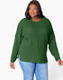A woman wearing a green ribbed knit long-sleeve top with a crew neck and side pockets, paired with blue jeans, against a plain white background.
