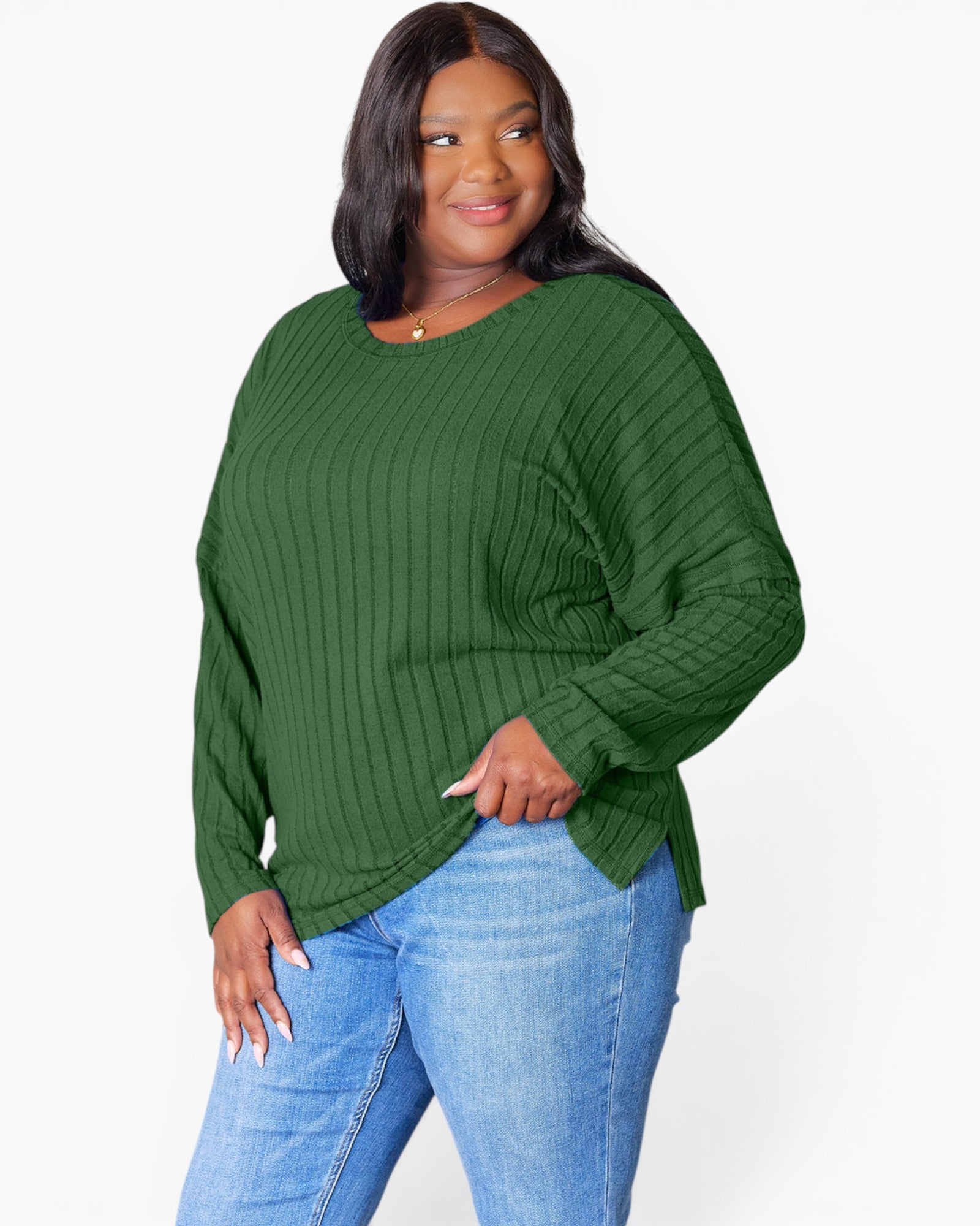 A woman wearing a dark green ribbed knit long-sleeve top and light blue jeans, standing against a plain white studio background.