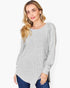A woman wearing a light gray ribbed knit long-sleeve tunic top with a high-low hem, paired with dark blue jeans, against a plain white background.