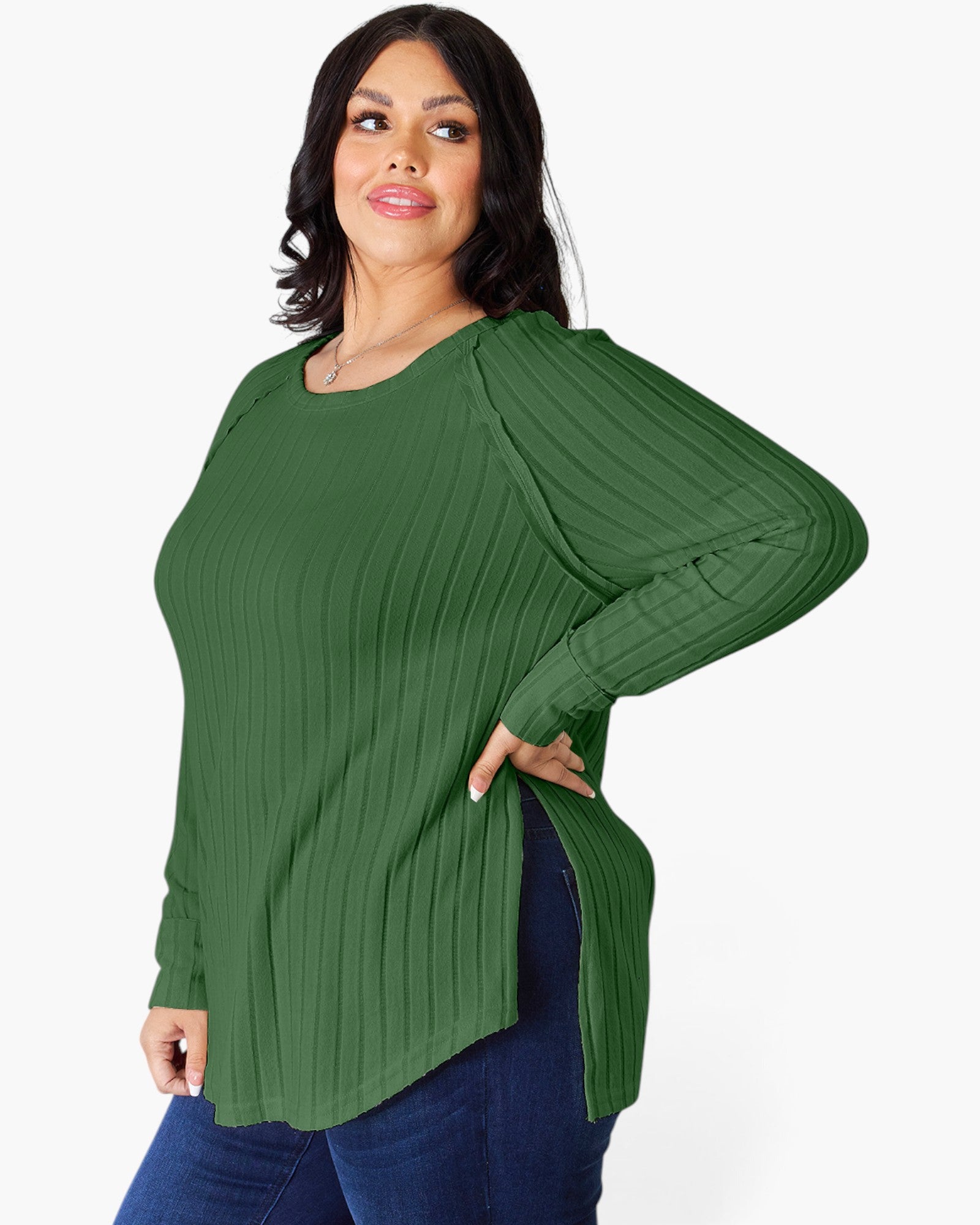 A woman modeling a green long-sleeve ribbed knit top with a high-low hem and side slits, paired with dark blue jeans, against a plain white background.