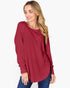 A woman wearing a long-sleeve, high-neck pleated top in a deep burgundy red, paired with blue jeans, against a plain white background.