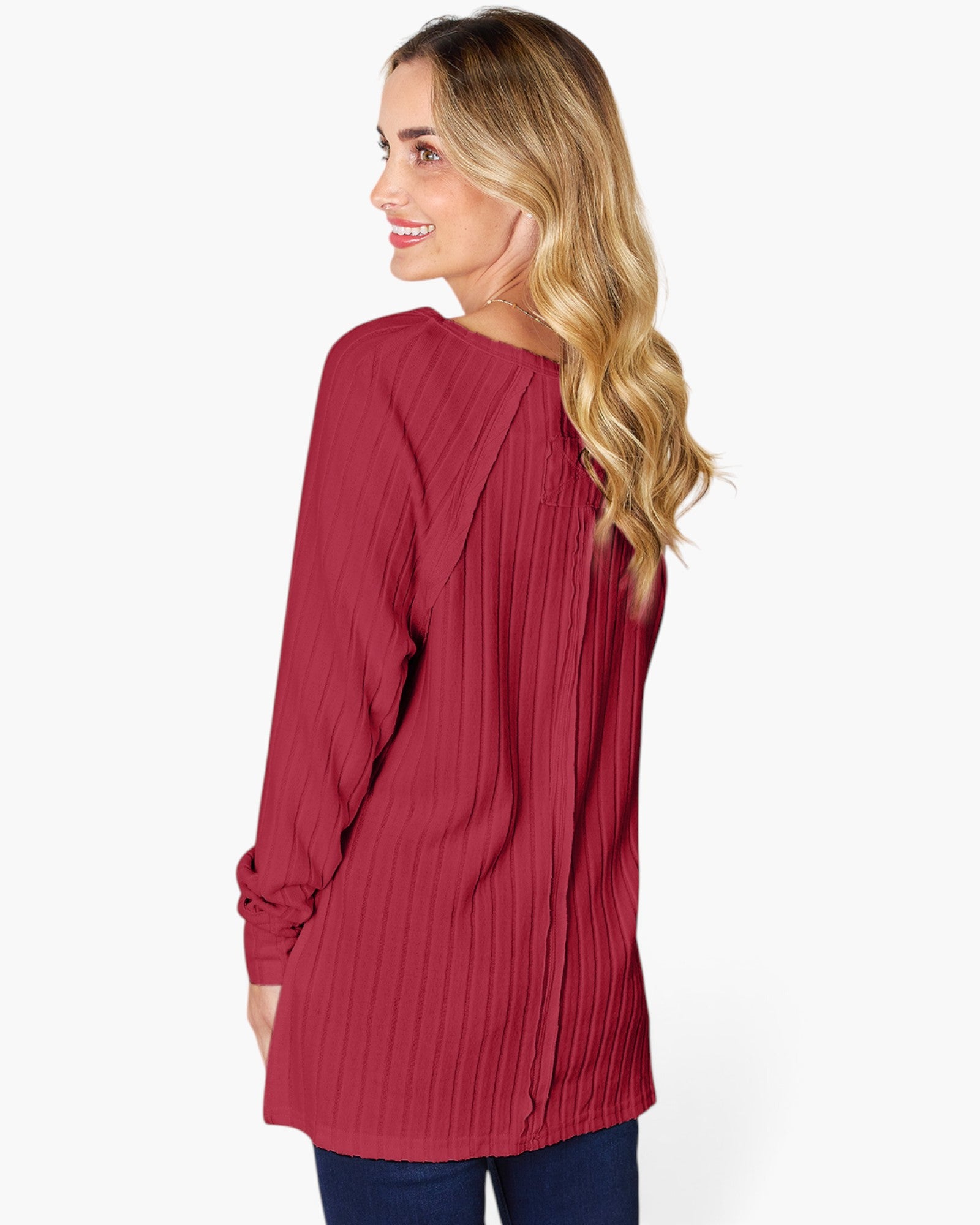 A woman with blonde wavy hair is shown from the back wearing a long-sleeve, pleated tunic top in a deep red or burgundy color, paired with dark blue jeans, against a plain white studio background.