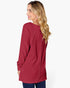 A woman with blonde wavy hair is shown from the back wearing a long-sleeve, pleated tunic top in a deep red or burgundy color, paired with dark blue jeans, against a plain white studio background.