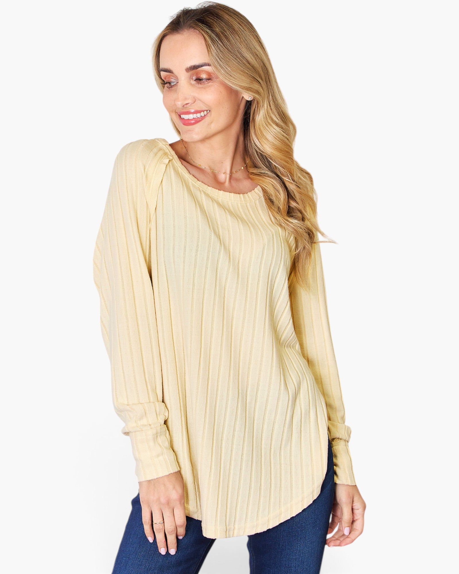 A woman wearing a pale yellow long-sleeve top with vertical pleats and blue jeans, photographed against a plain light gray studio background.