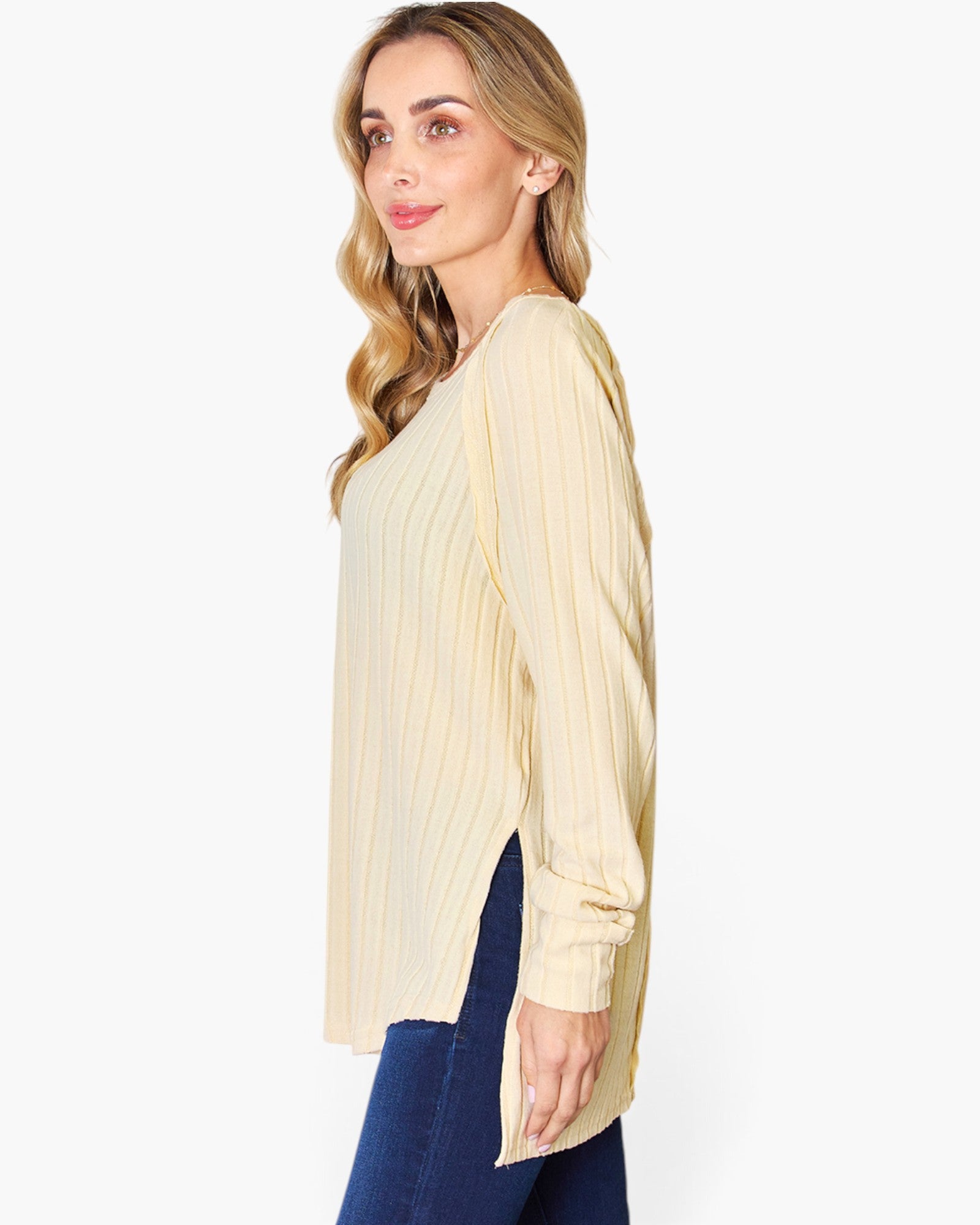 A woman wearing a cream-colored, long-sleeve ribbed knit top with side slits, paired with dark blue jeans, shown from a side profile against a plain white background.