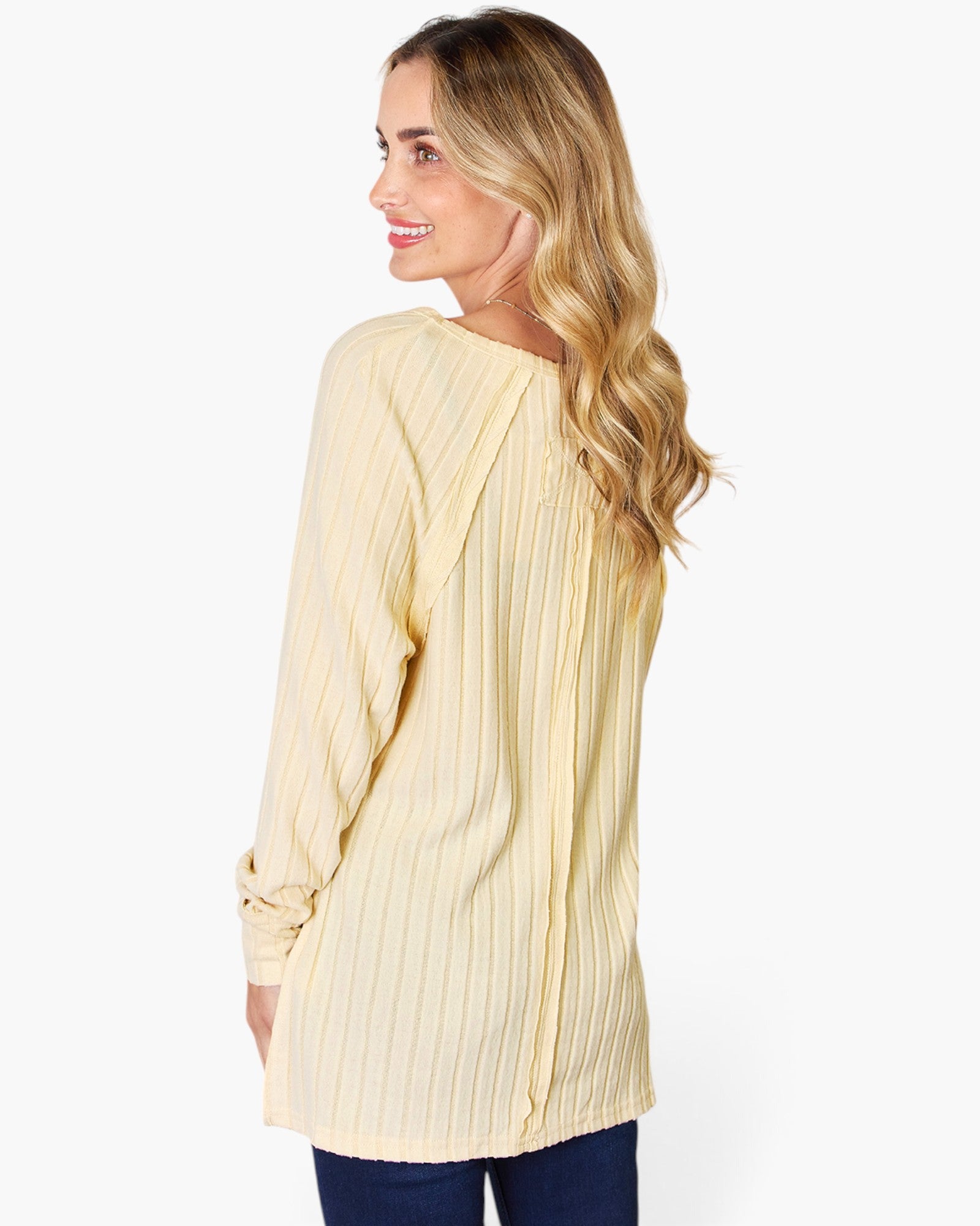 A woman shown from the back wearing a pale yellow long-sleeve top with vertical pleats, paired with dark blue jeans, against a plain white background.