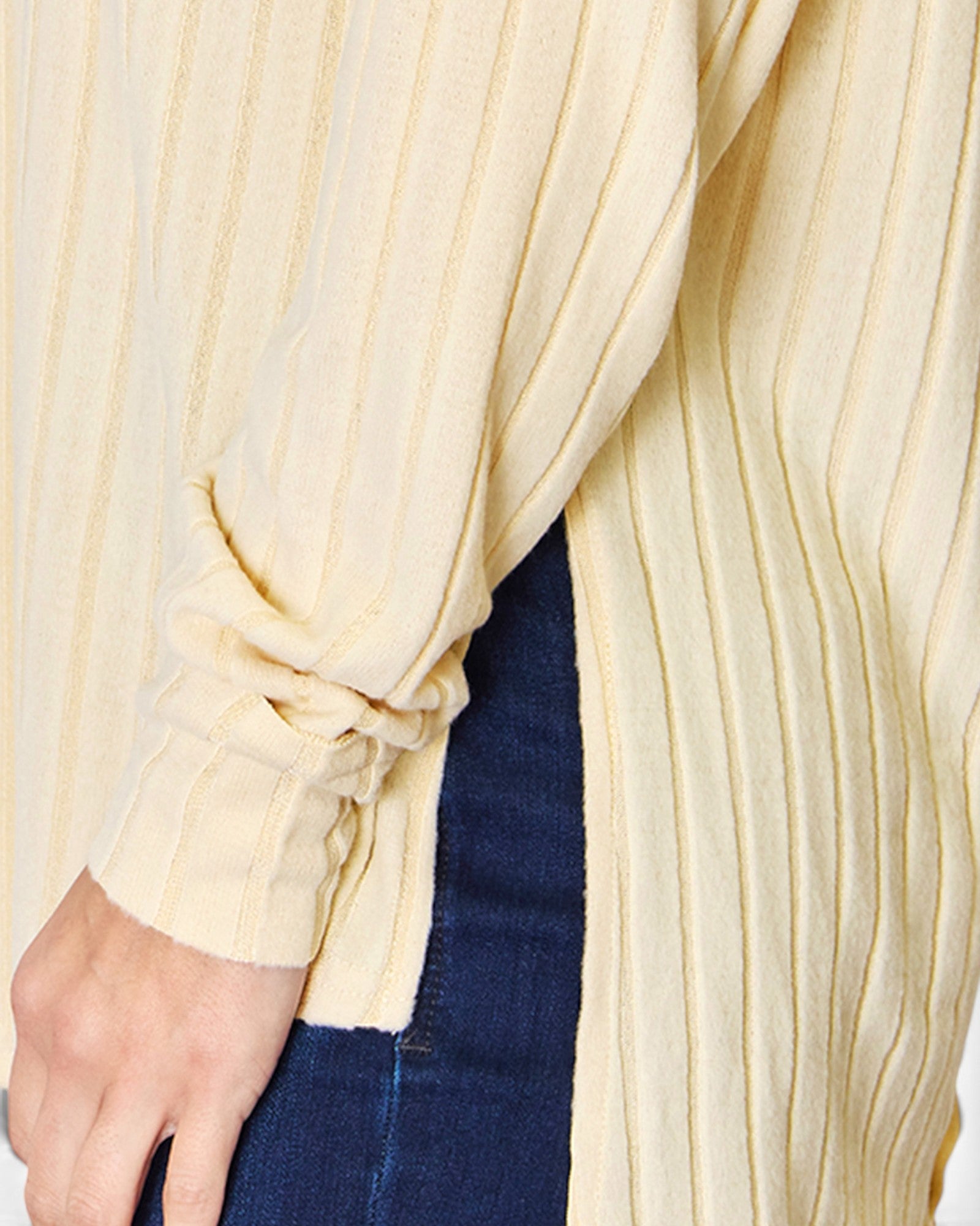 Close-up of a cream ribbed knit cardigan with a rolled-up sleeve and side slit, worn over dark blue jeans.