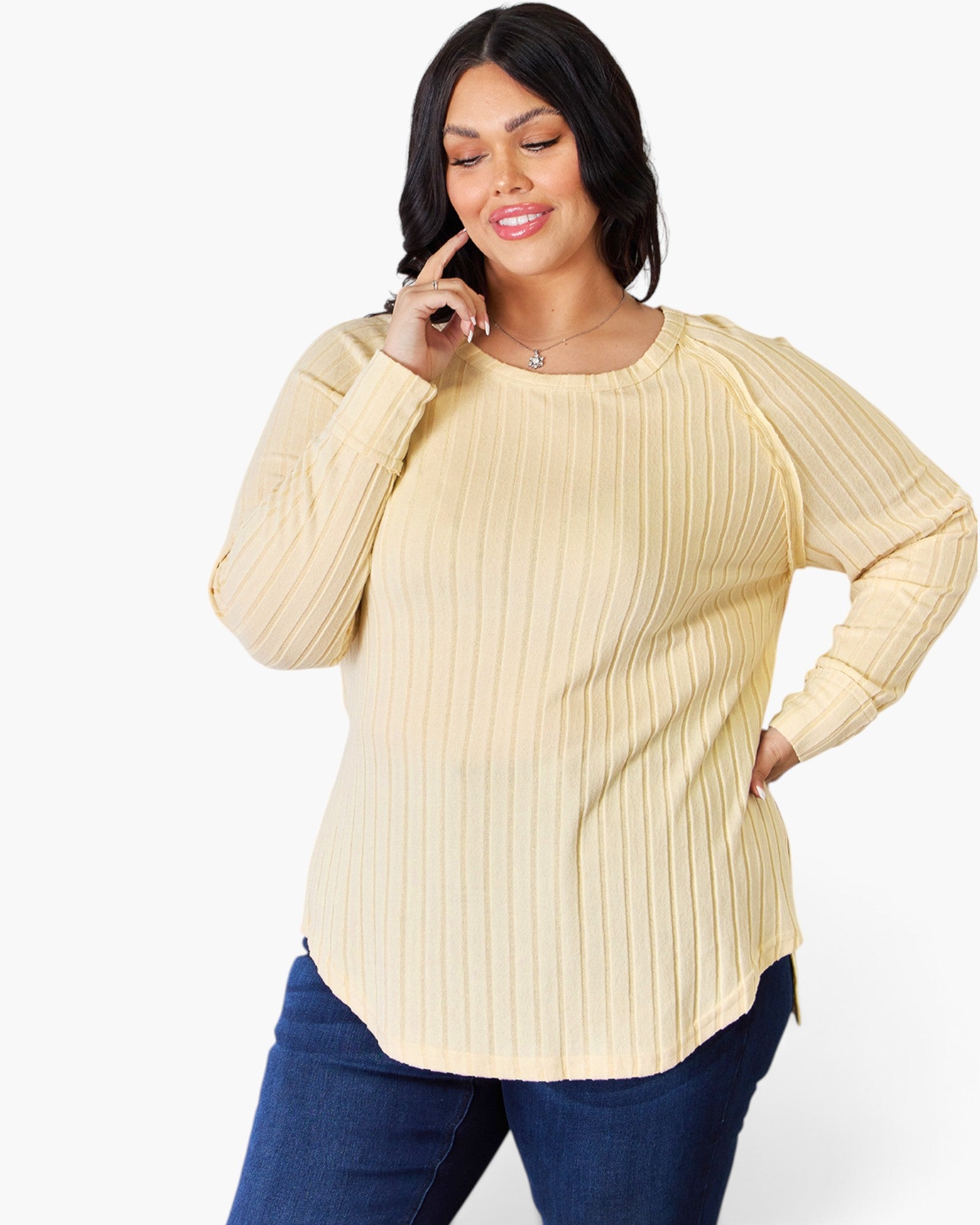 A woman wearing a cream-colored ribbed long-sleeve top and dark blue jeans, posing against a solid white background.