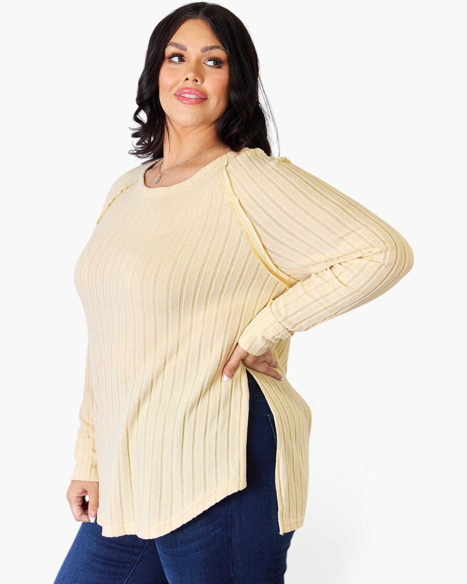 A woman modeling a cream ribbed knit long-sleeve top with unique draped shoulder details and a side slit, paired with dark blue jeans against a plain white background.