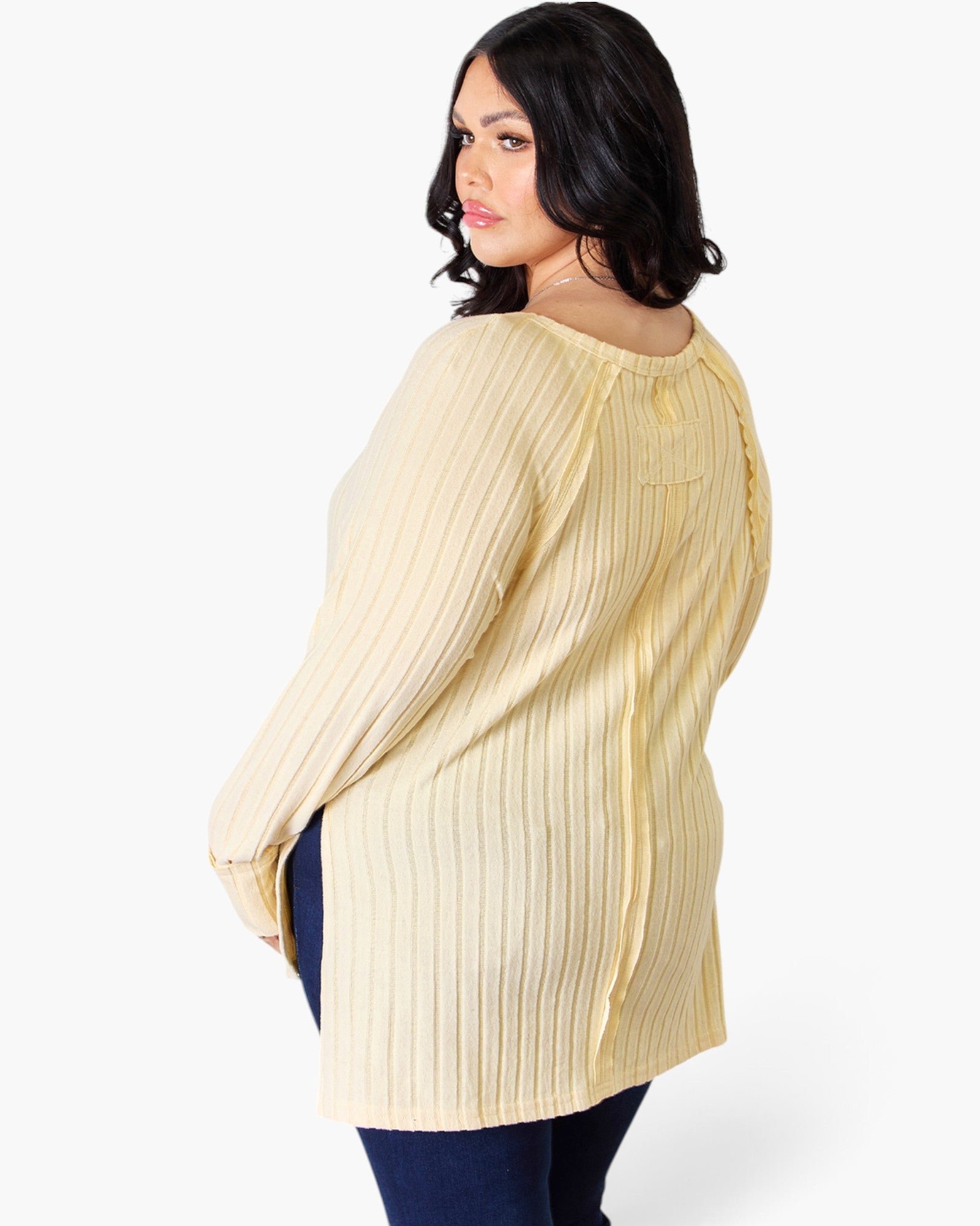 A woman wearing a cream-colored ribbed knit tunic sweater with a back knot detail, shown from a three-quarter back angle against a light gray studio background.