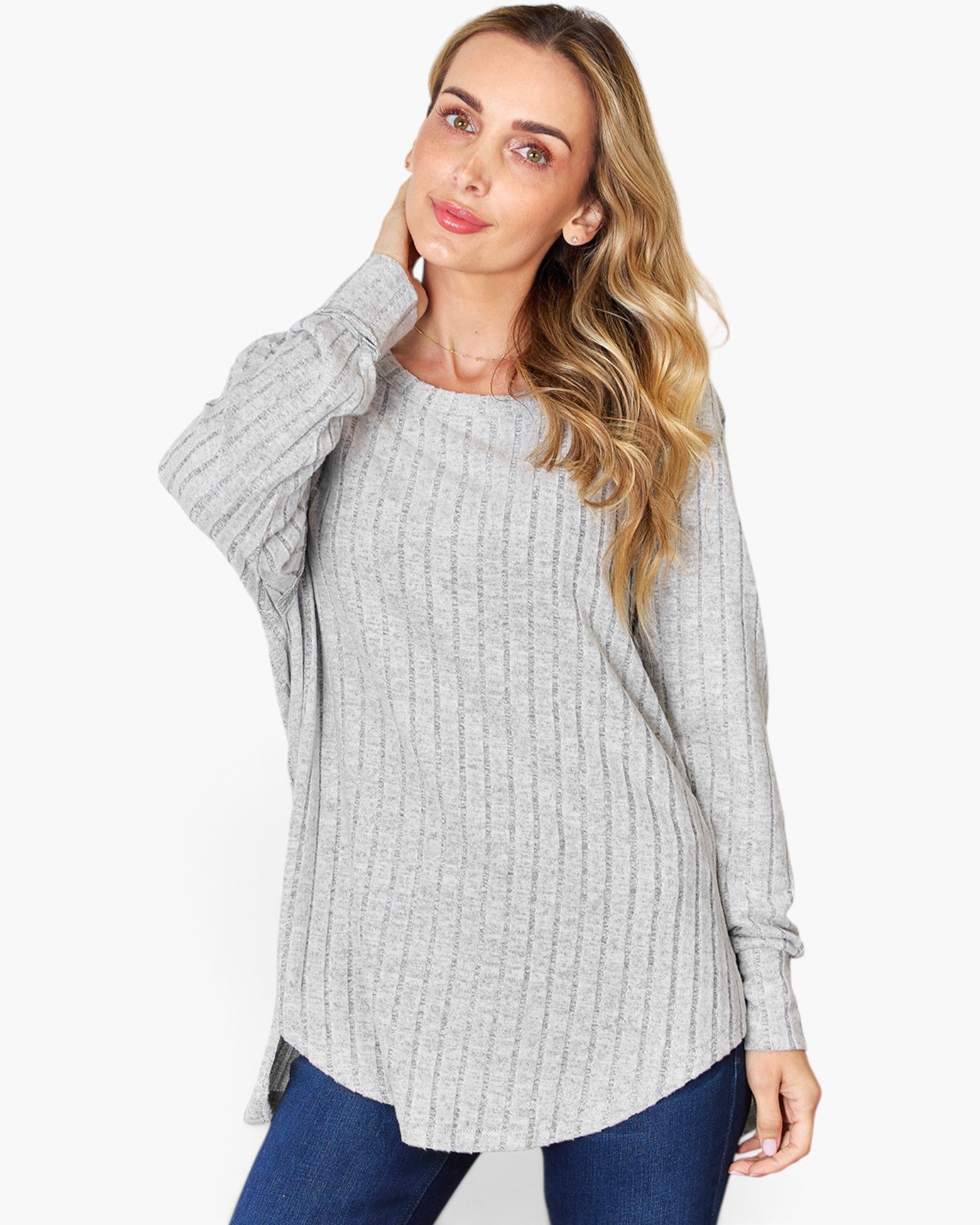 A woman wearing a light gray ribbed knit long-sleeve tunic top with blue jeans, posing against a solid white studio background.
