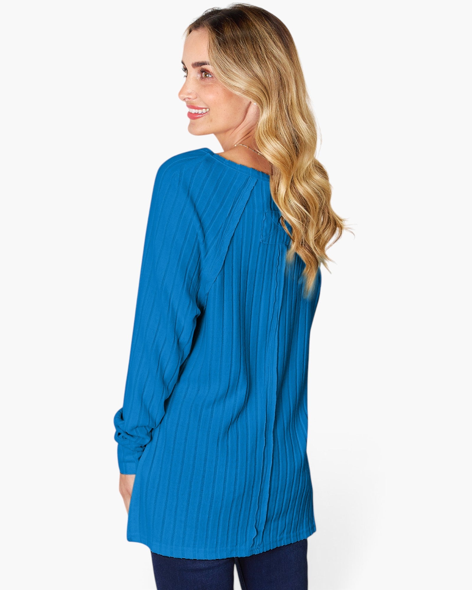 A woman with blonde wavy hair is shown from the back and side, wearing a bright blue long-sleeve top with vertical pleats and dark navy pants, against a plain white studio background.