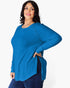 A woman modeling a bright blue ribbed knit long-sleeve top with a round neckline and side slit, paired with dark blue jeans against a light gray studio background.