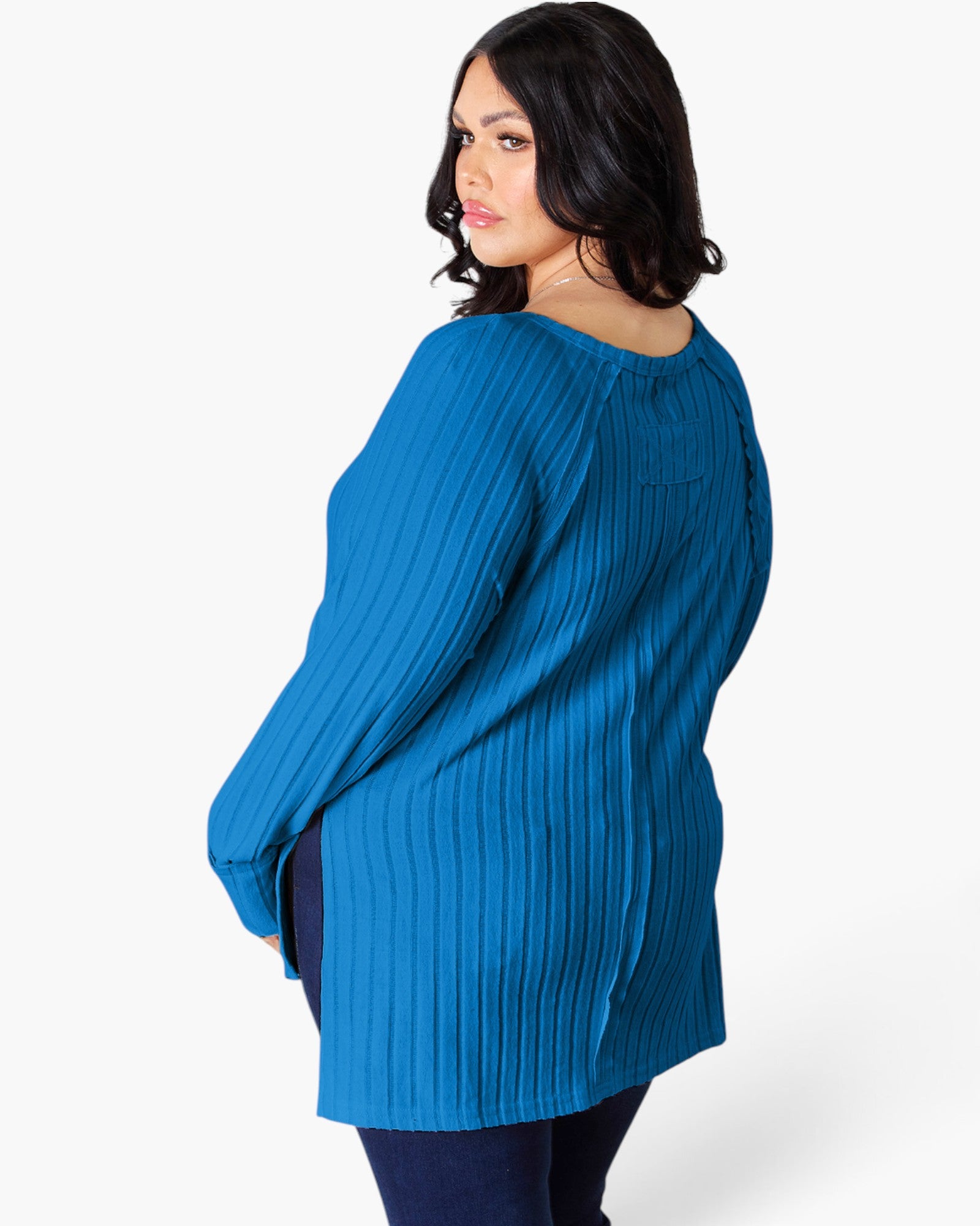 A woman wearing a bright blue ribbed knit long-sleeve tunic top, shown from a three-quarters back view against a light gray studio background.